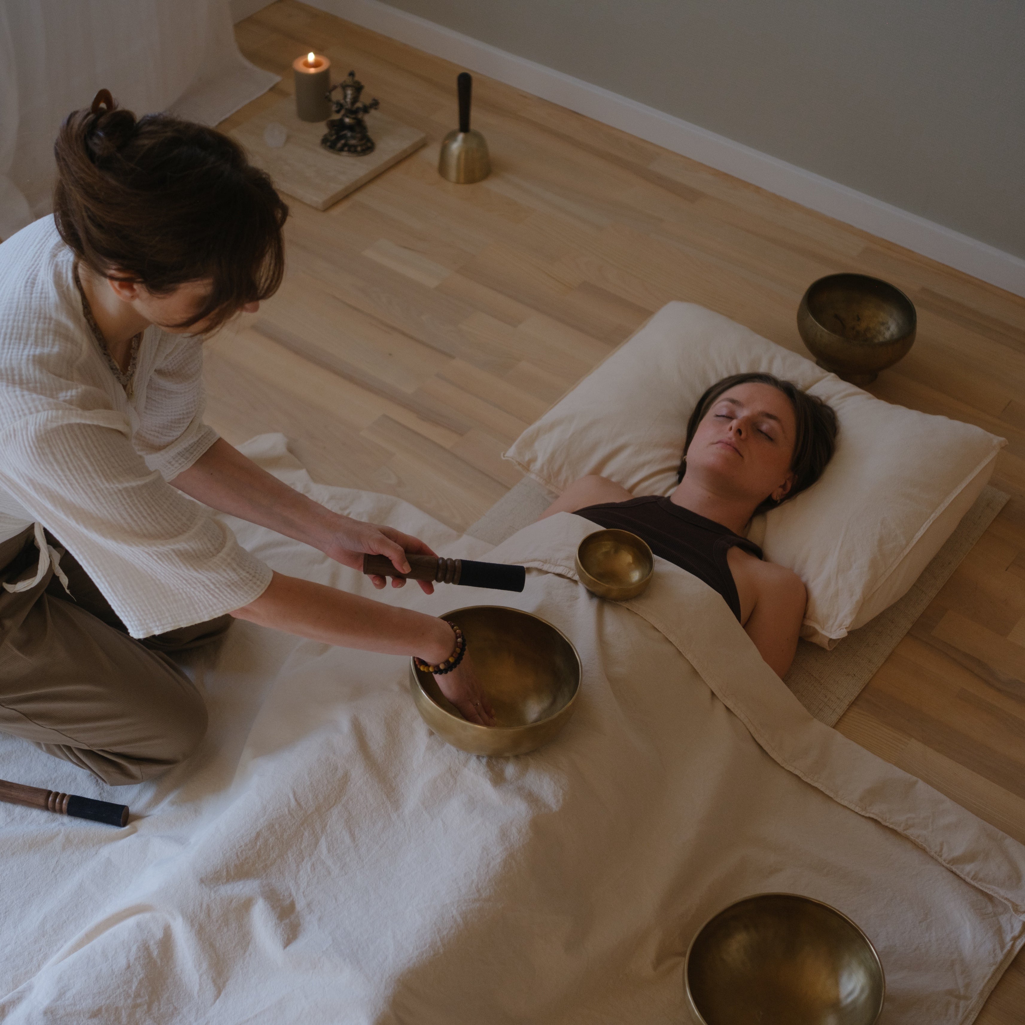 Person receiving a sound bath treatment with another person with dim singing bowls in a serene room.