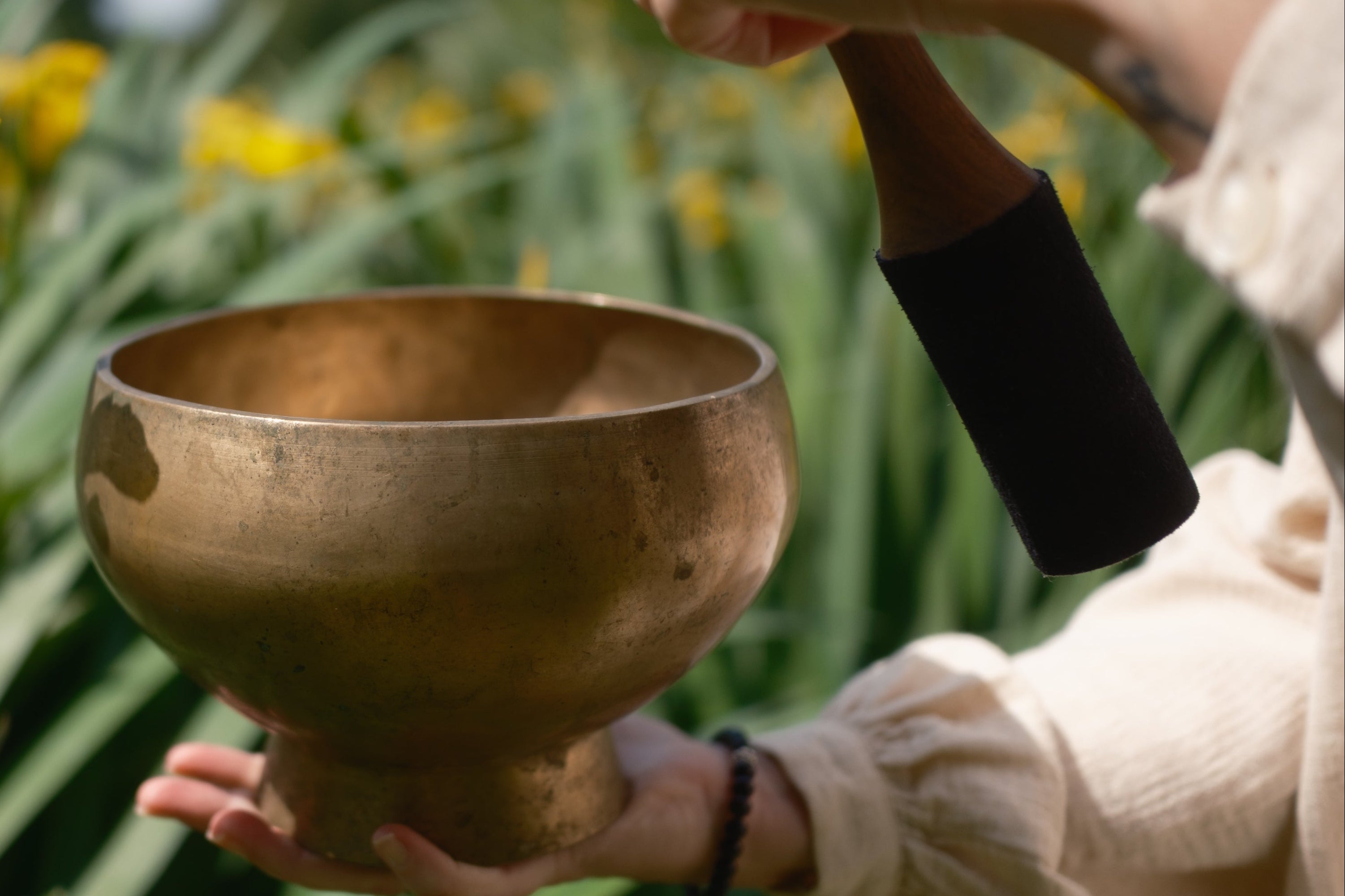Person holding a antique pedestal singing bowl outdoors with yellow flowers and greenery in the background