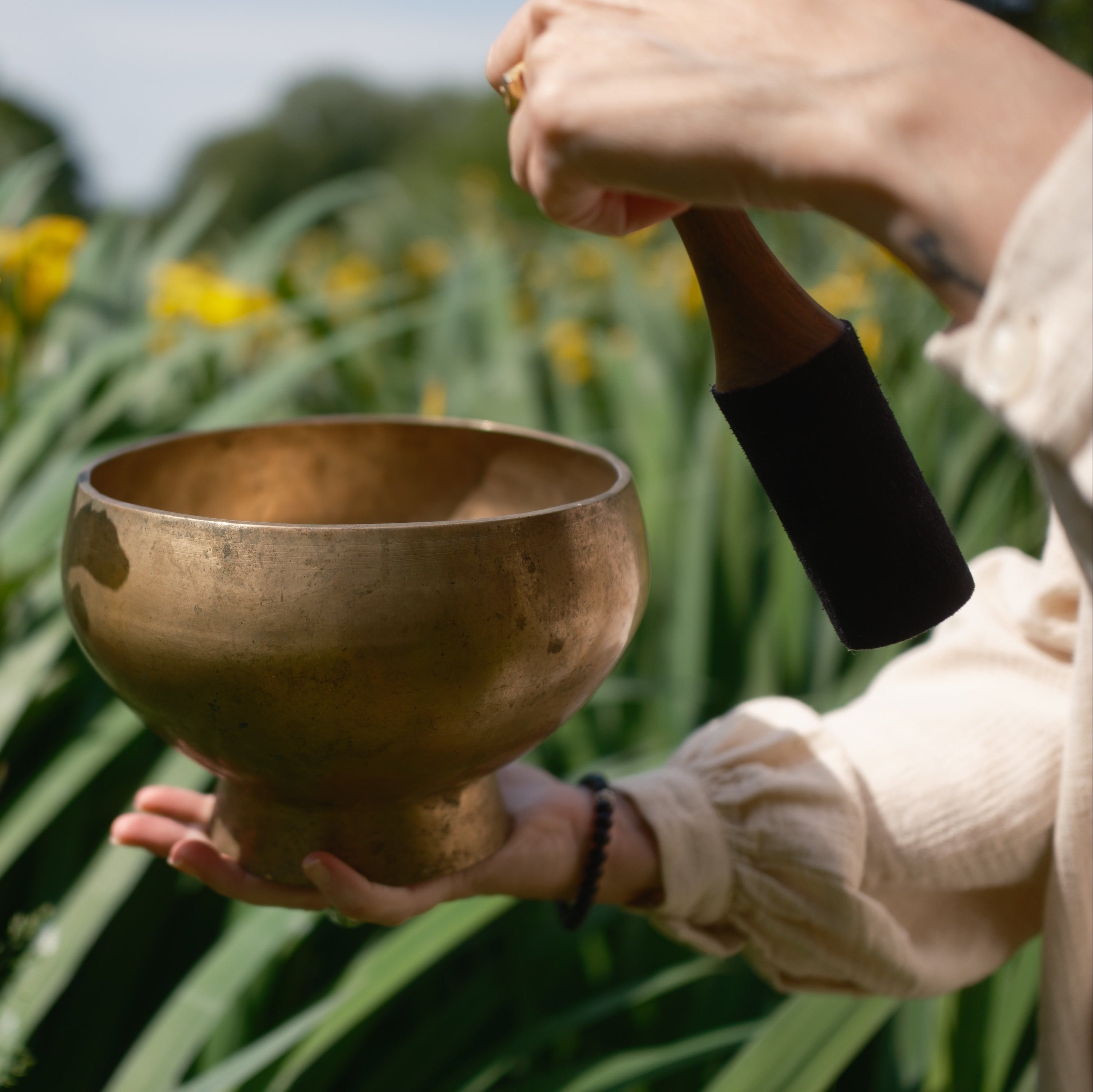 Person holding a antique pedestal singing bowl outdoors with yellow flowers and greenery in the background