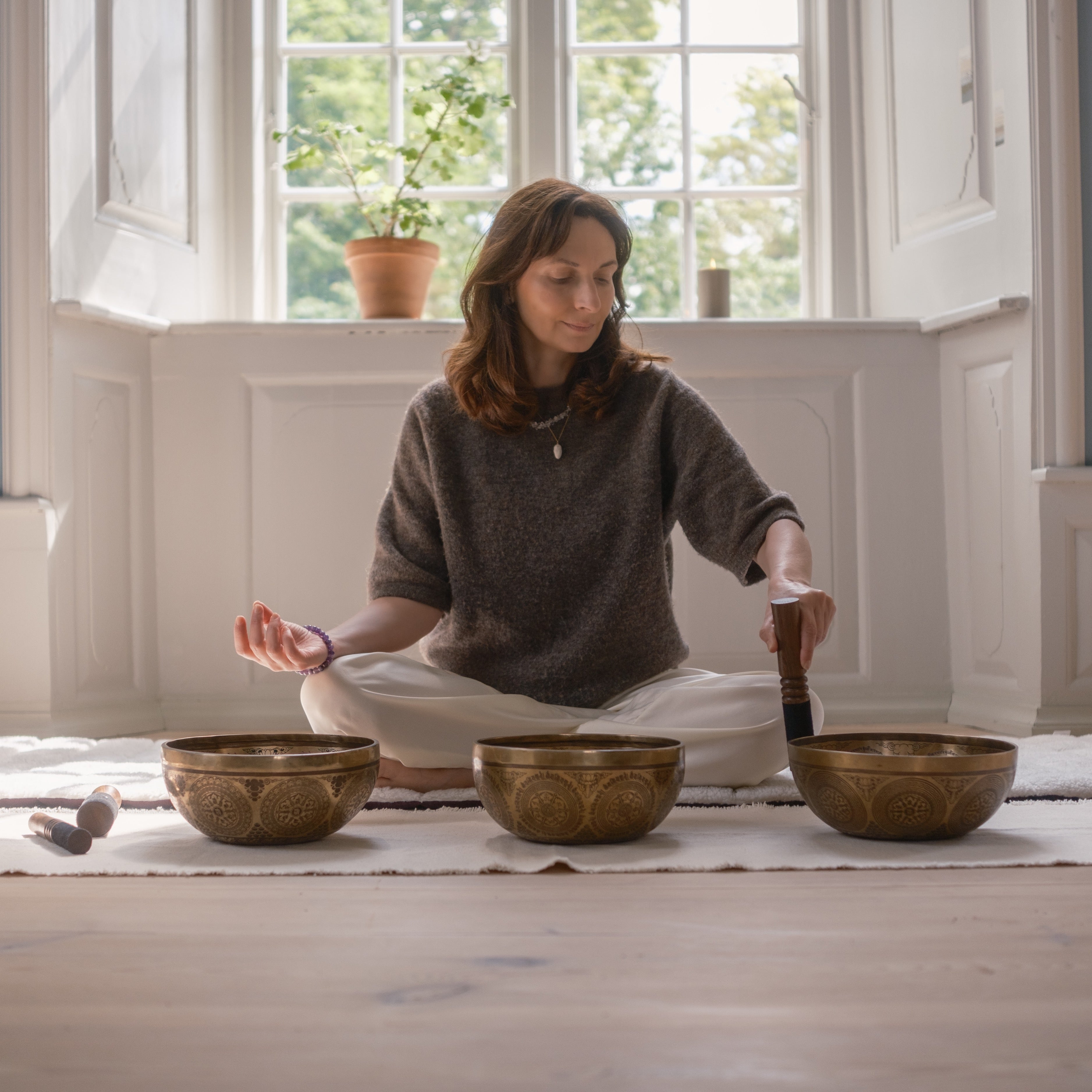 Woman sitting in a meditative pose with singing bowls in a room with large windows.