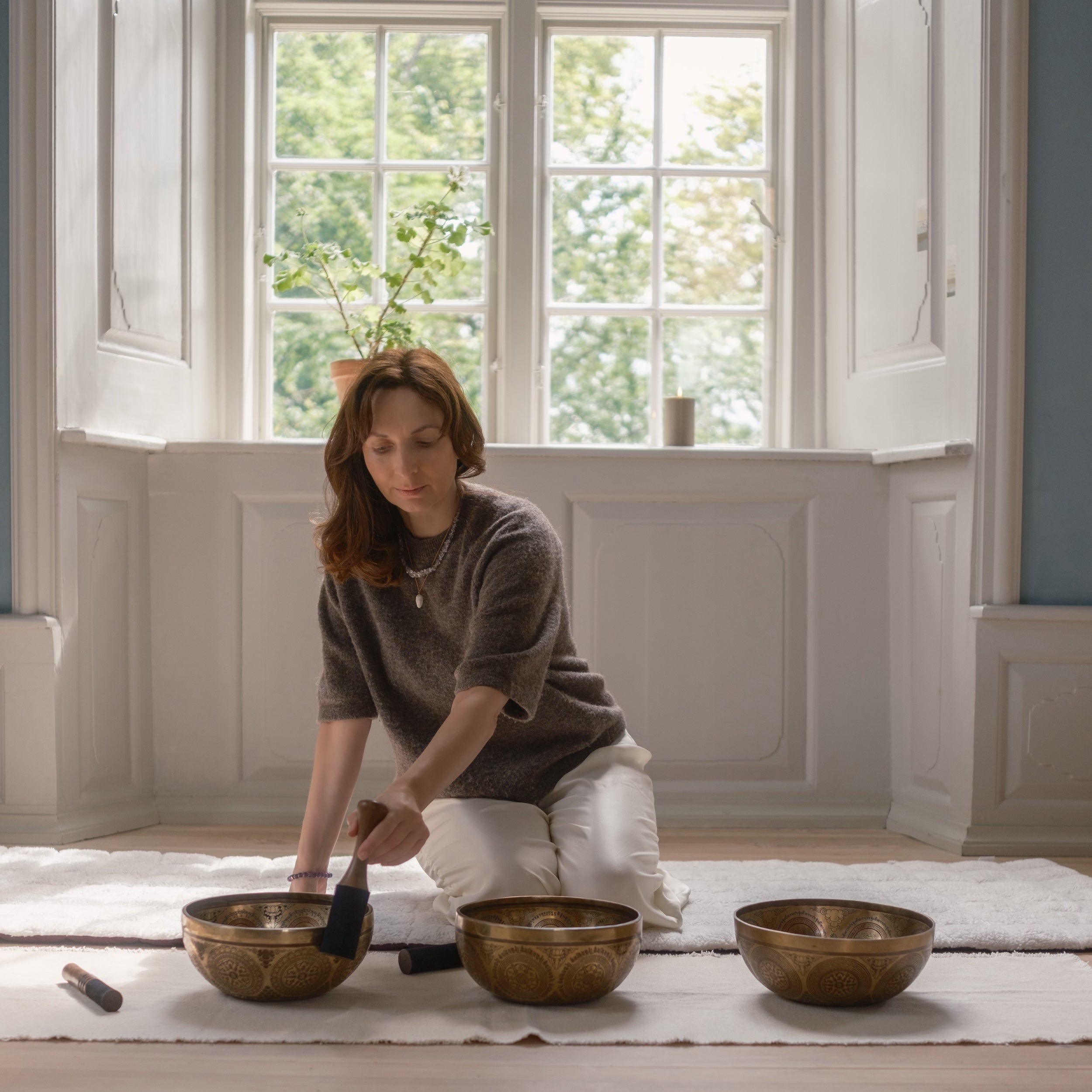 Woman playing on a set of three bronze singing bowls in a room with large windows.