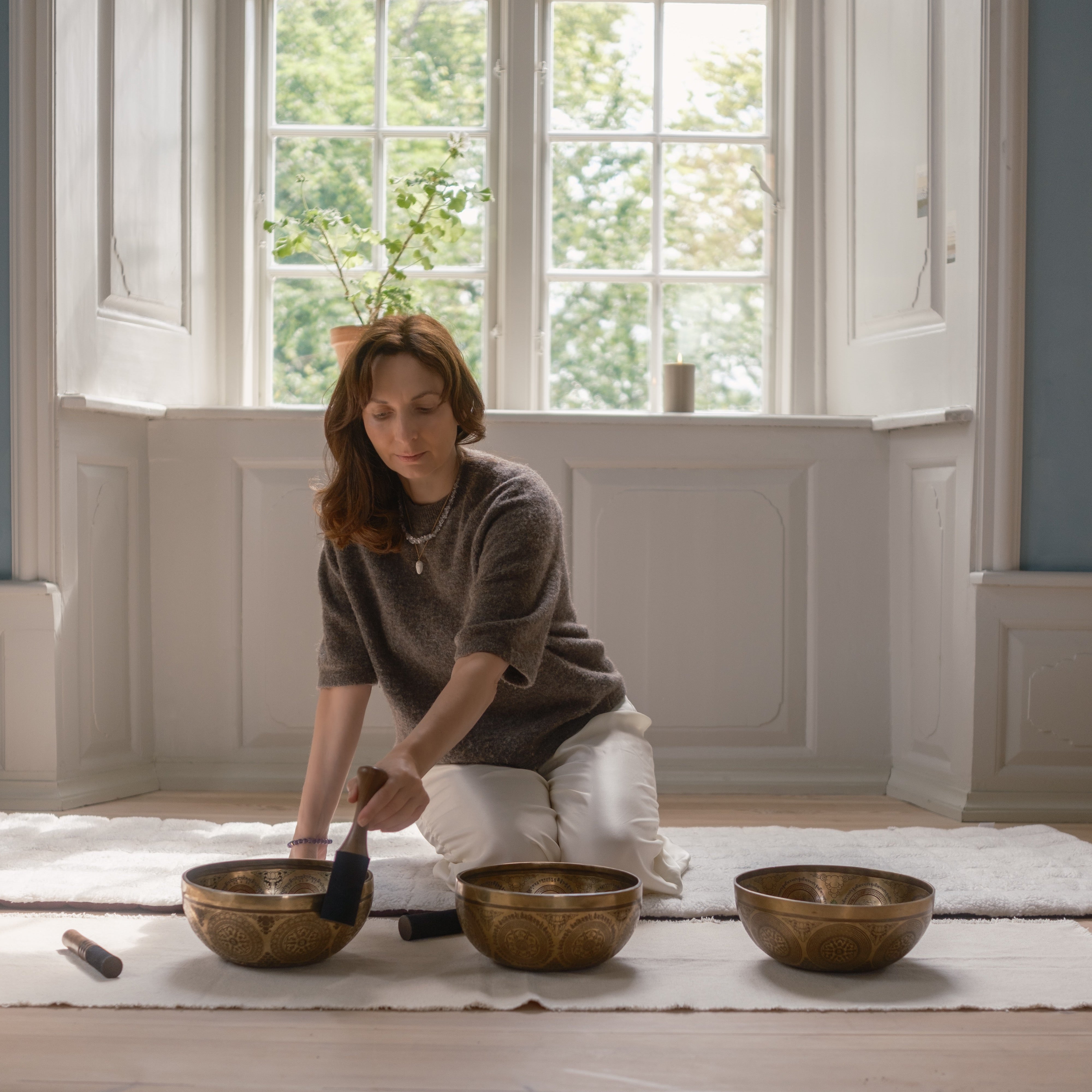 Woman sitting on the floor with three bronze singing bowls in a room with large windows.