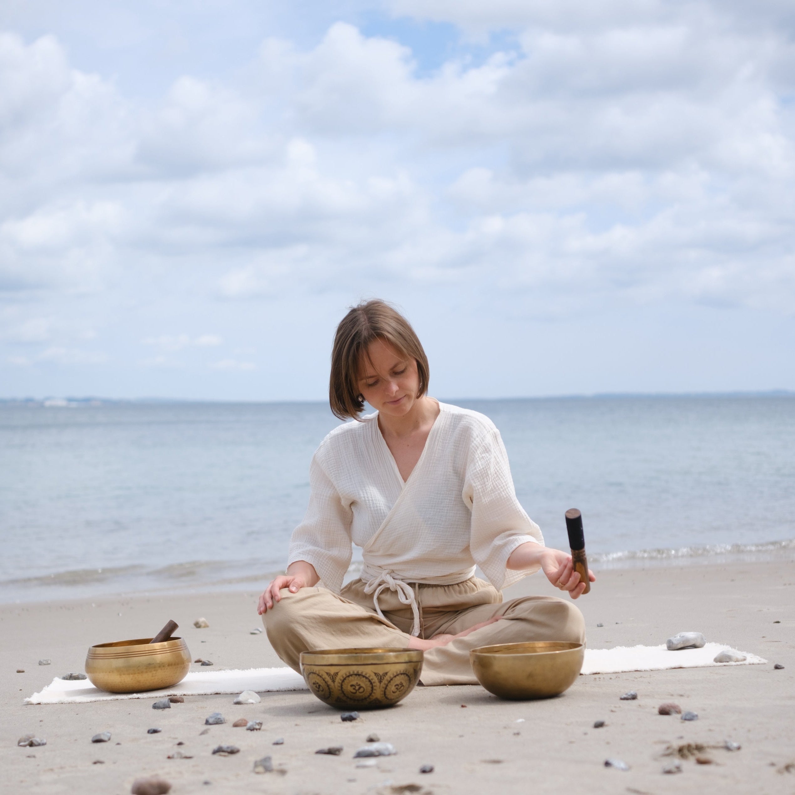Woman playing singing bowls on a beach with ocean and sky in the background