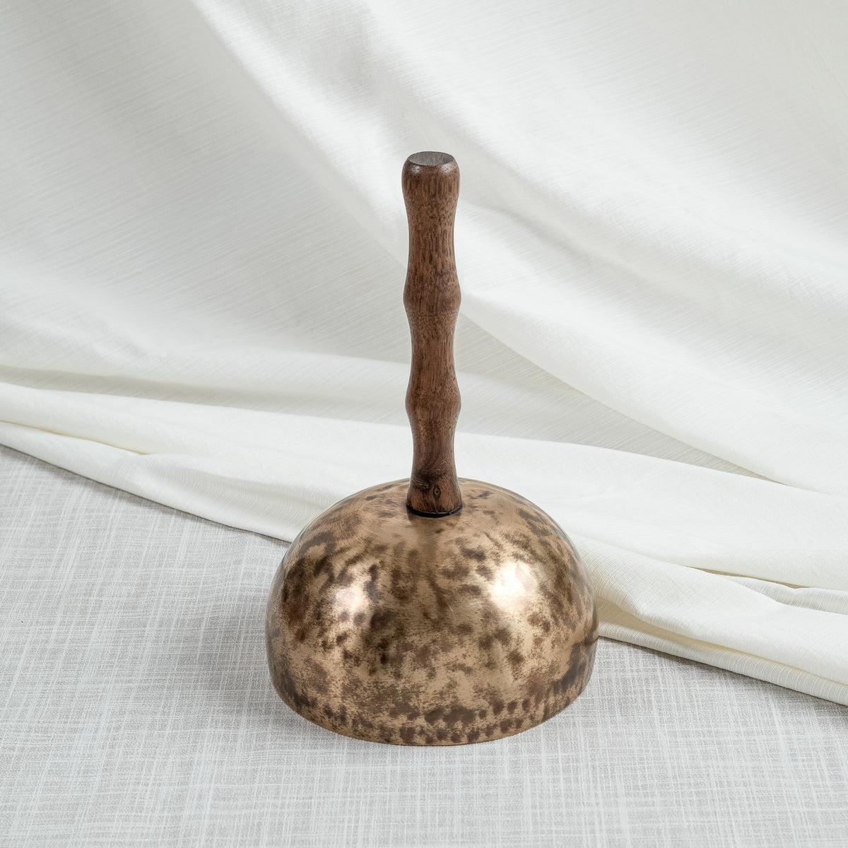 A hand-hammered singing bowls with handle with a dark wooden handle, photographed on a light textured surface with draped white fabric in the background.