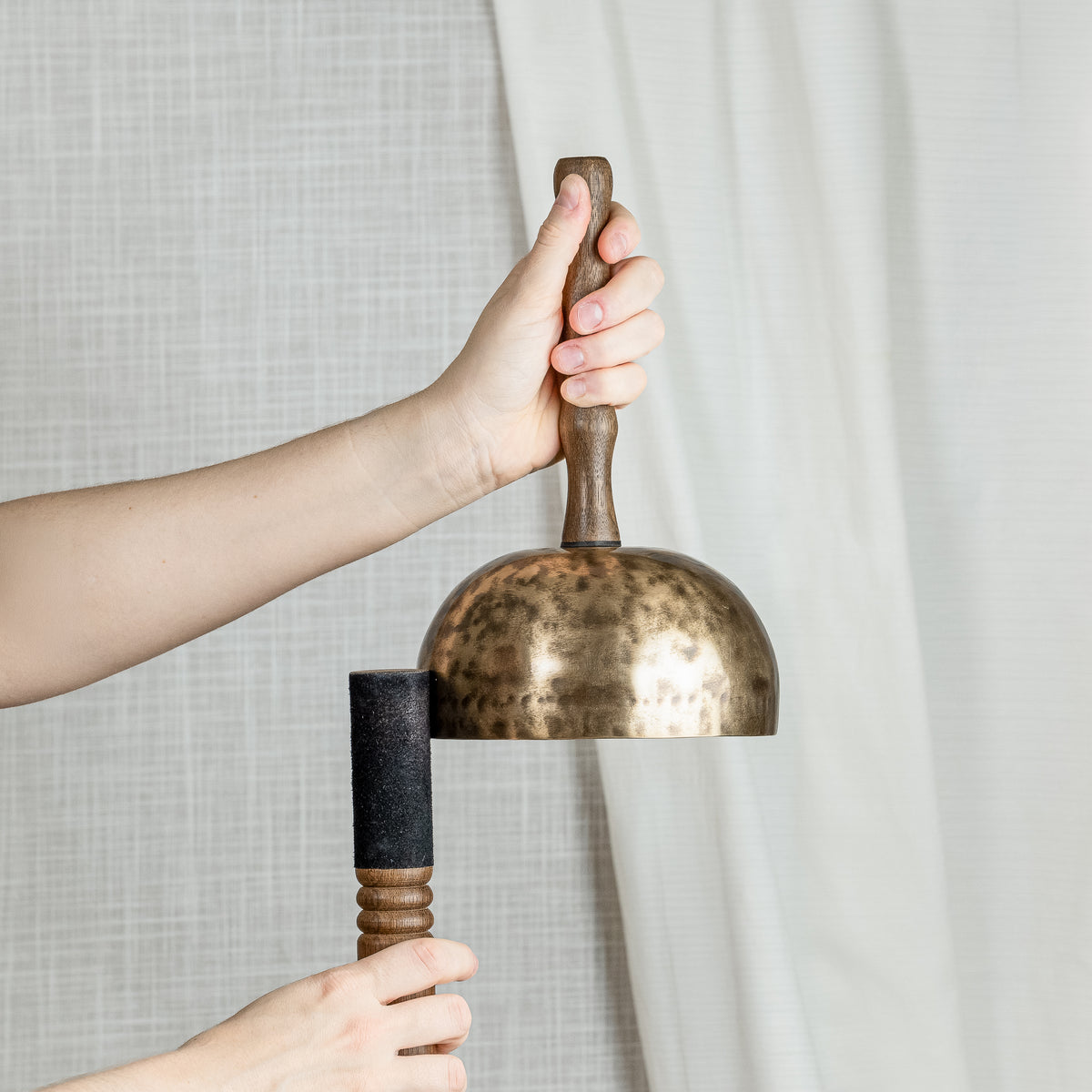 A person holds a bronze singing bowl with a wooden handle and a black striker against a neutral background of textured gray fabric and white drapes.