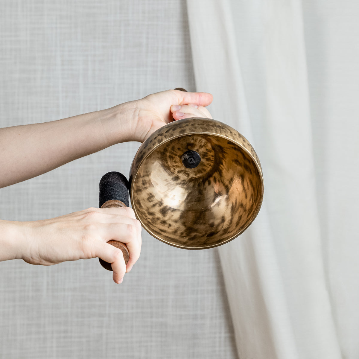 A bronze singing bowl with a hammered finish and wooden handle, held by hands against a neutral light gray curtain background.