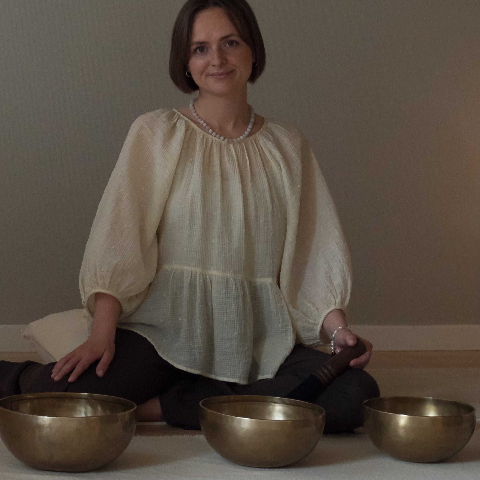 Woman sitting with three dim singing bowls on a mat against a plain background