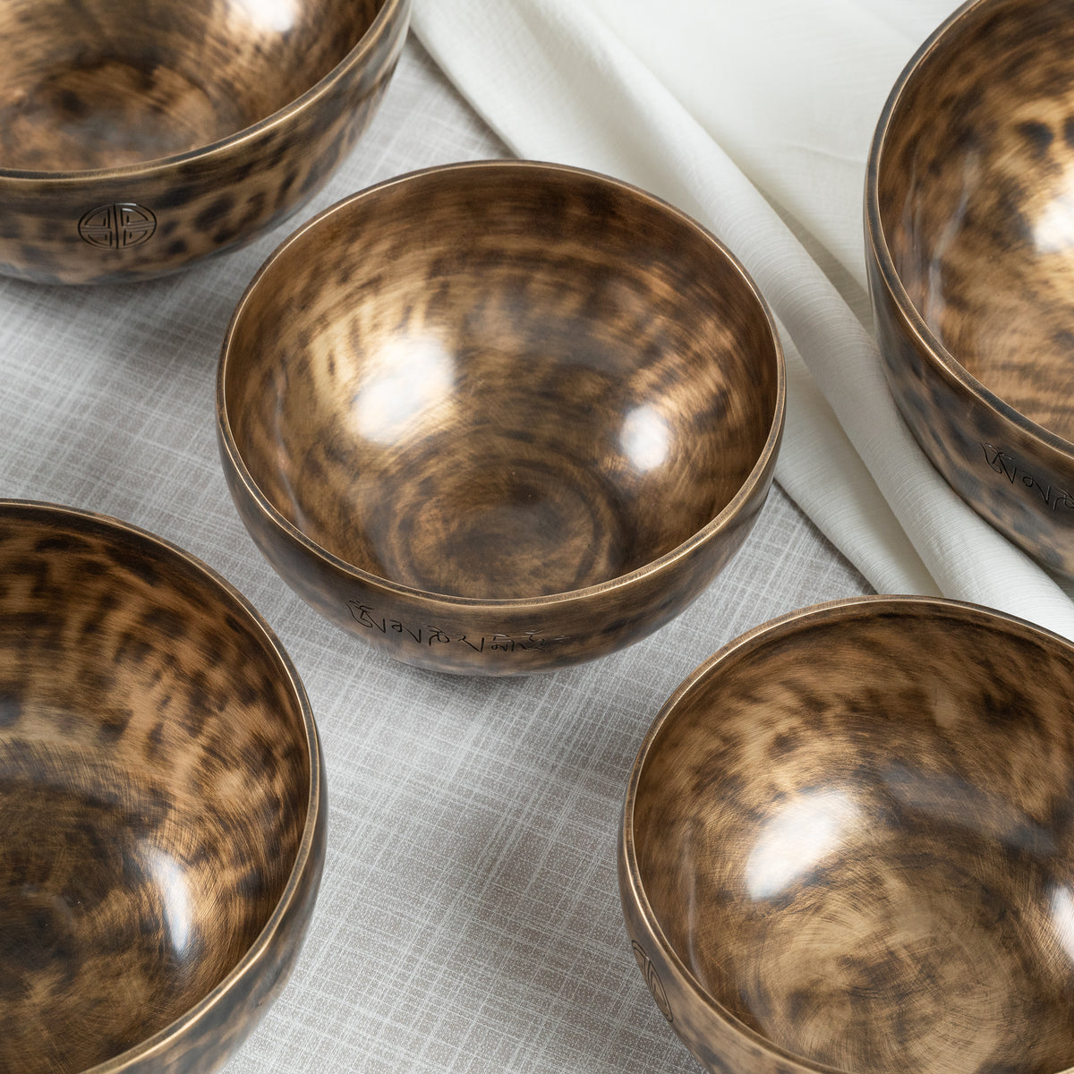 Four metallic singing bowls with a hammered bronze-brown finish, arranged on a light gray textured cloth with a white fabric accent.