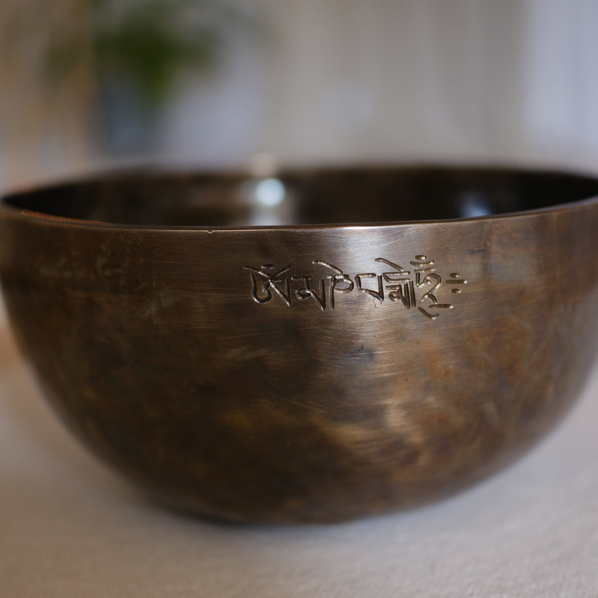 A close-up of a bronze singing bowl with engraved Tibetan script, resting on a light beige surface with a softly blurred background.
