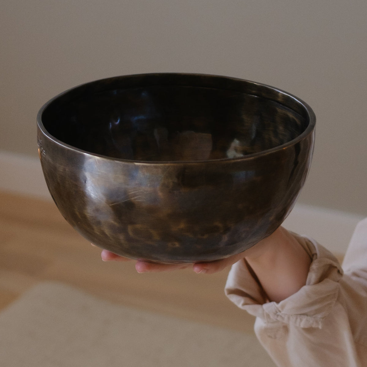 A person in a beige long-sleeve shirt holds a dark, hammered lunar energy singing bowl against a soft beige background.