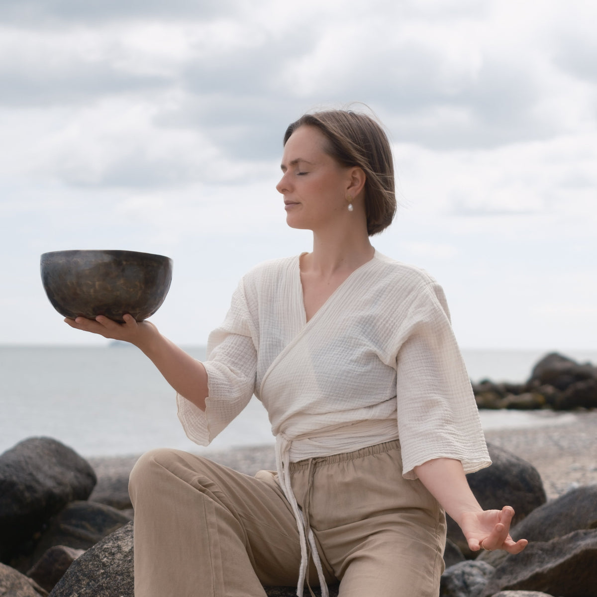 A woman sits on rocks by the ocean holding a dark hand-hammered singing bowl, wearing a cream wrap top and beige pants under a cloudy sky.