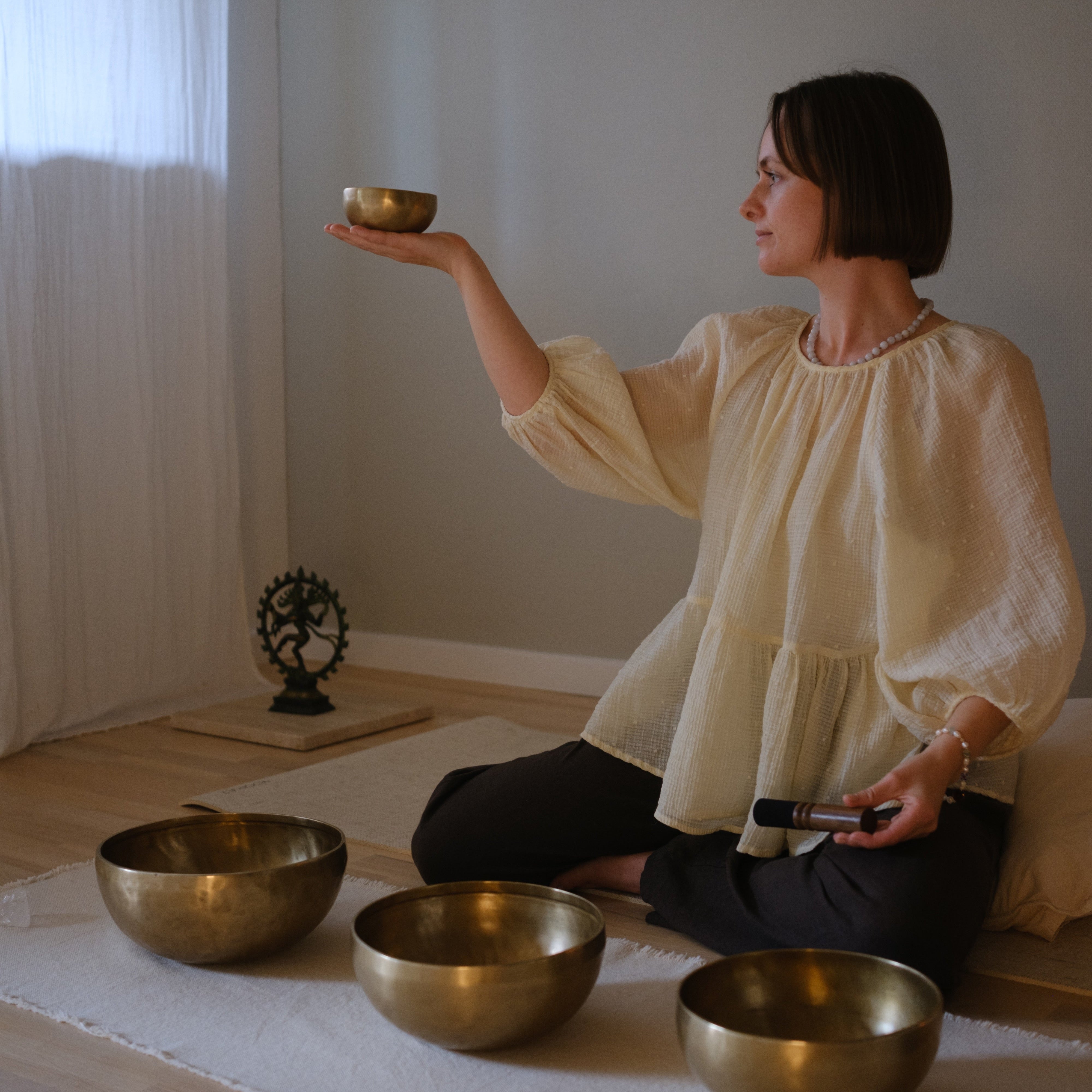 Woman sitting on a mat with small dim singing bowls in a room with white walls and a curtain.