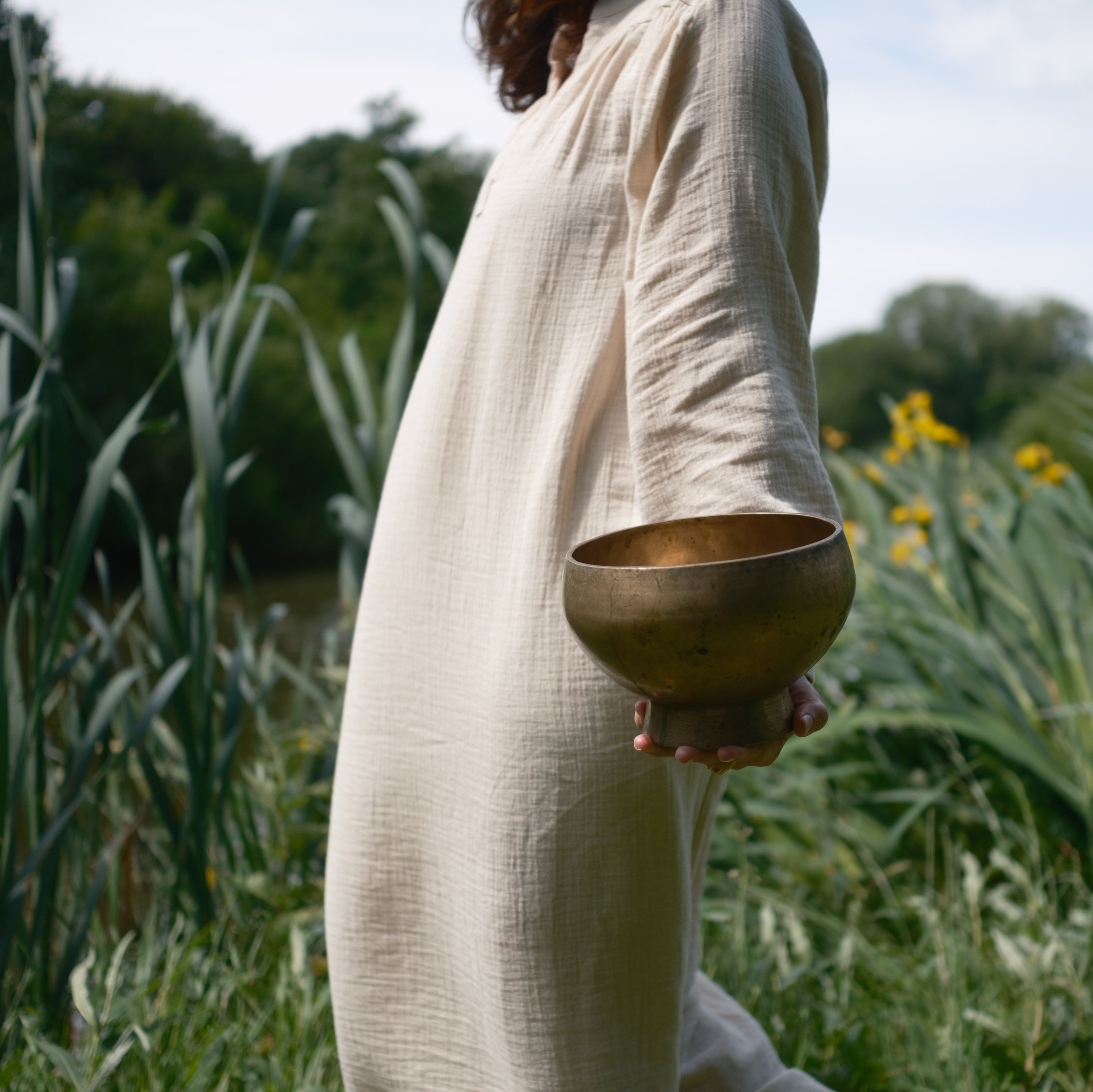 Person holding a antique pedestal singing bowl in a natural setting with greenery and flowers.