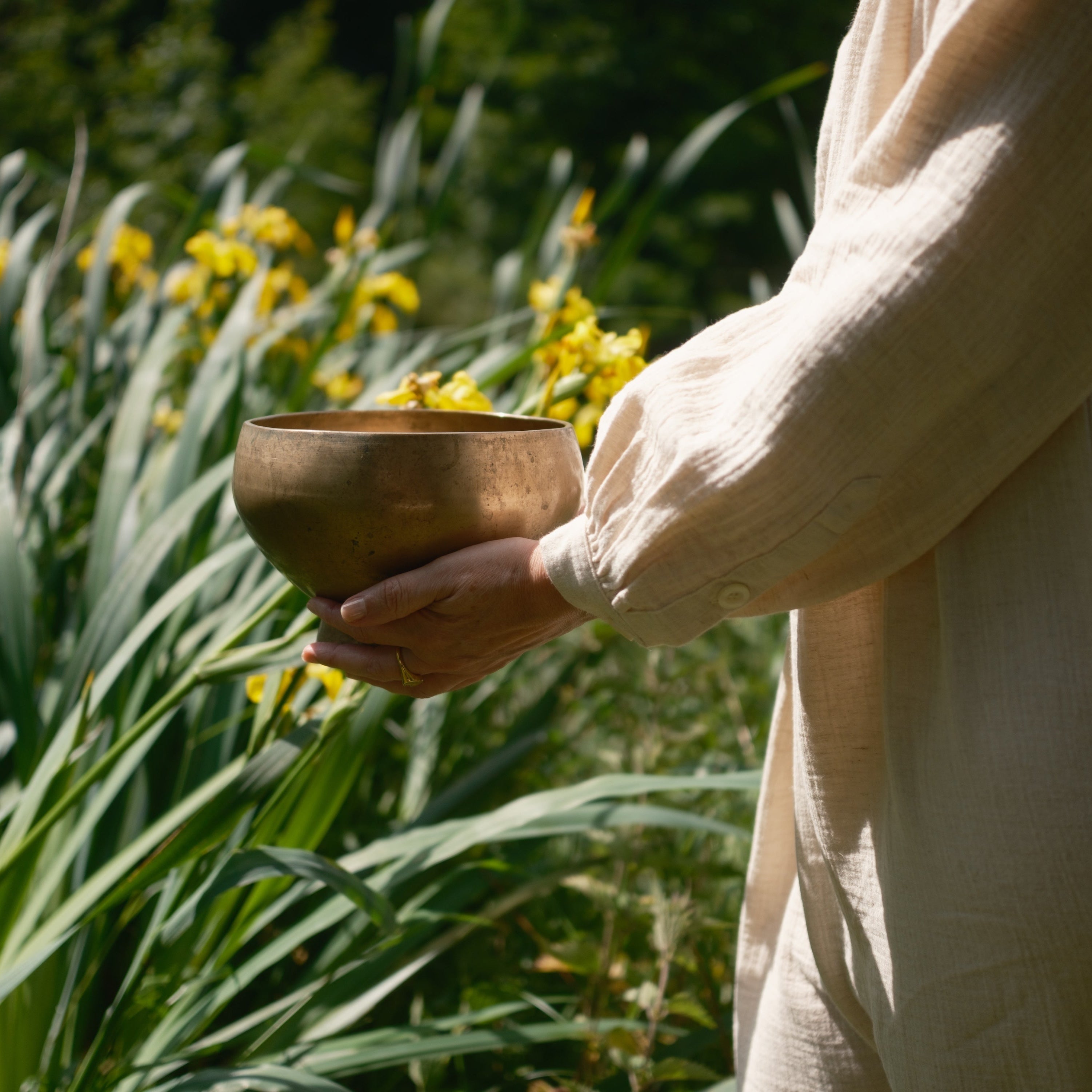 Person, der holder en antik piedestal-syngeskål i naturlige omgivelser med grønne planter og gule blomster.
