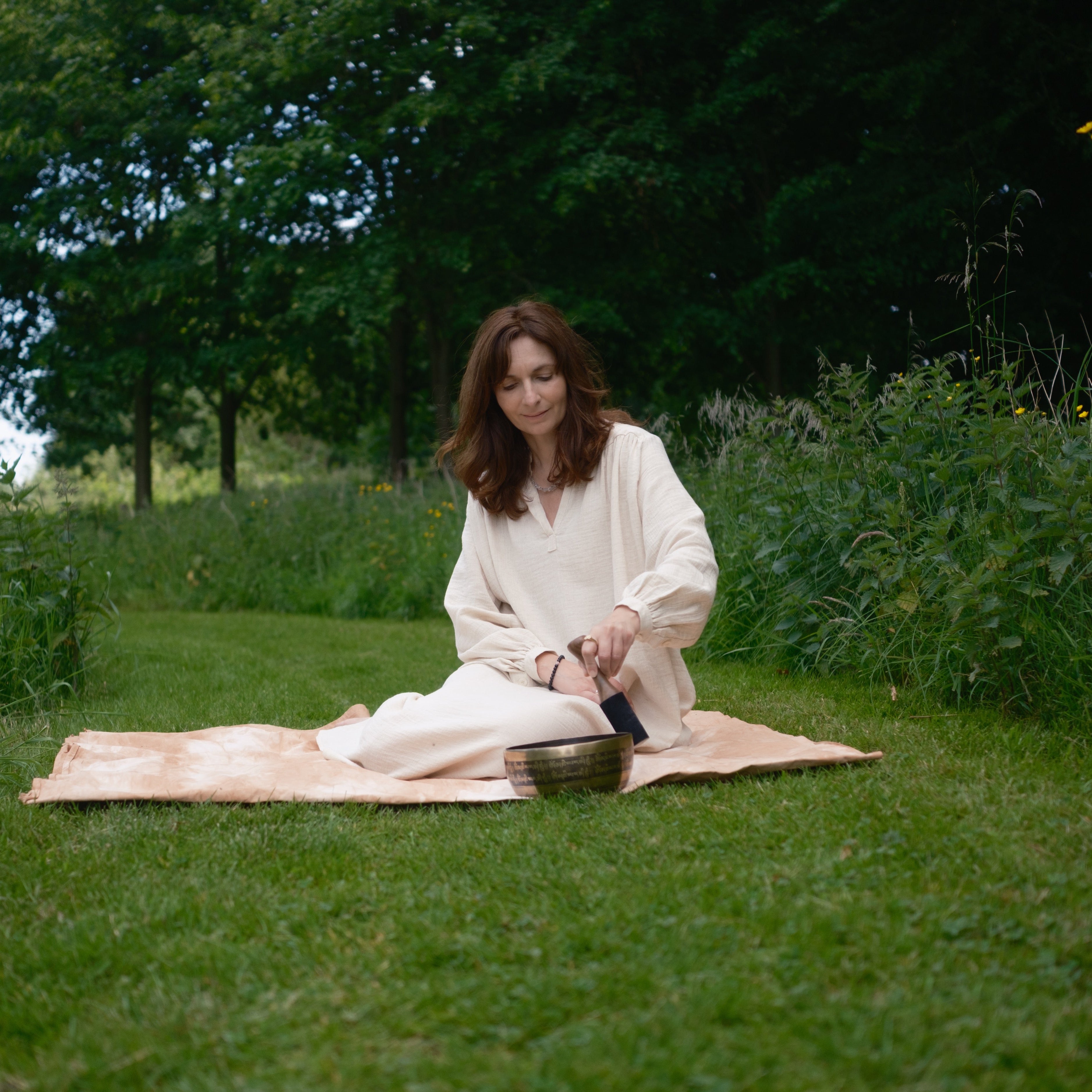 Woman in a white dress sitting on a blanket in a grassy field with trees in the background, playing a singing bowl