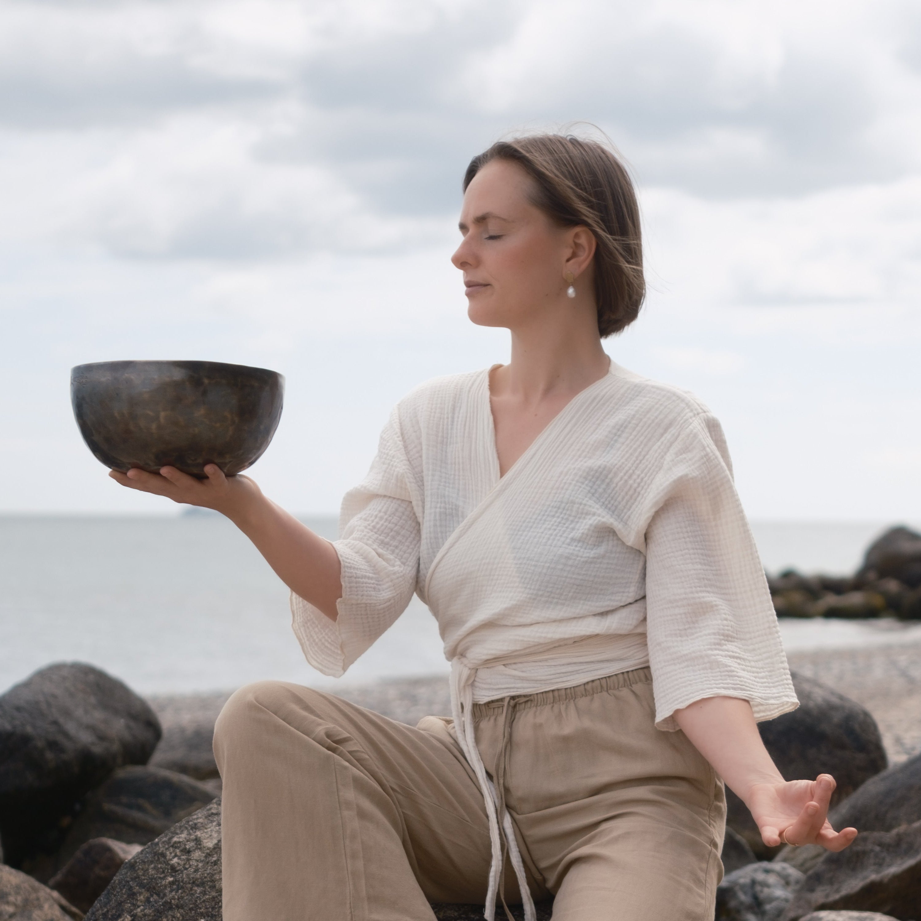Woman holding a lunar energy singing bowl on a rocky beach with ocean and sky in the background