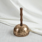 A hand-hammered singing bowls with handle with a dark wooden handle, photographed on a light textured surface with draped white fabric in the background.
