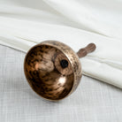 A hand-hammered singing bowl with handle with a dark wooden handle, photographed on a light textured fabric surface with draped white fabric in the background.