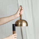 A person holds a bronze singing bowl with a wooden handle and a black striker against a neutral background of textured gray fabric and white drapes.