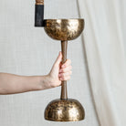 A hand holds a double sided singing bowl with handle, with a wooden striker against a neutral light gray fabric backdrop.