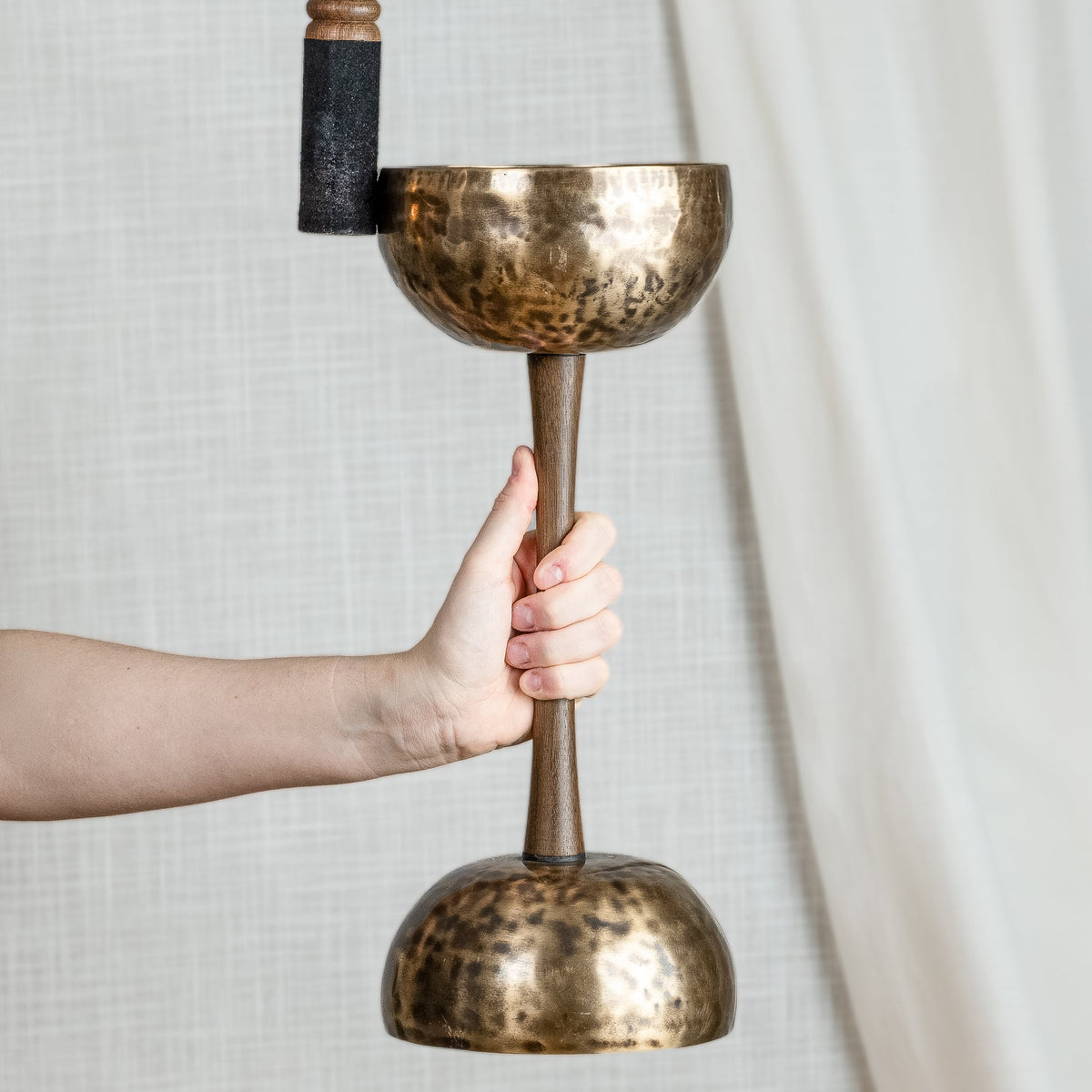 A hand holds a double sided singing bowl with handle, with a wooden striker against a neutral light gray fabric backdrop.