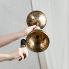 A double sided singing bowl with a wooden handle and black wrapped striker, held by hands against a neutral light gray textured wall and white curtain background.