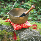 A antique singing bowl with a wooden striker rests on a red and gold patterned cushion placed on a mossy rock in an outdoor garden setting.