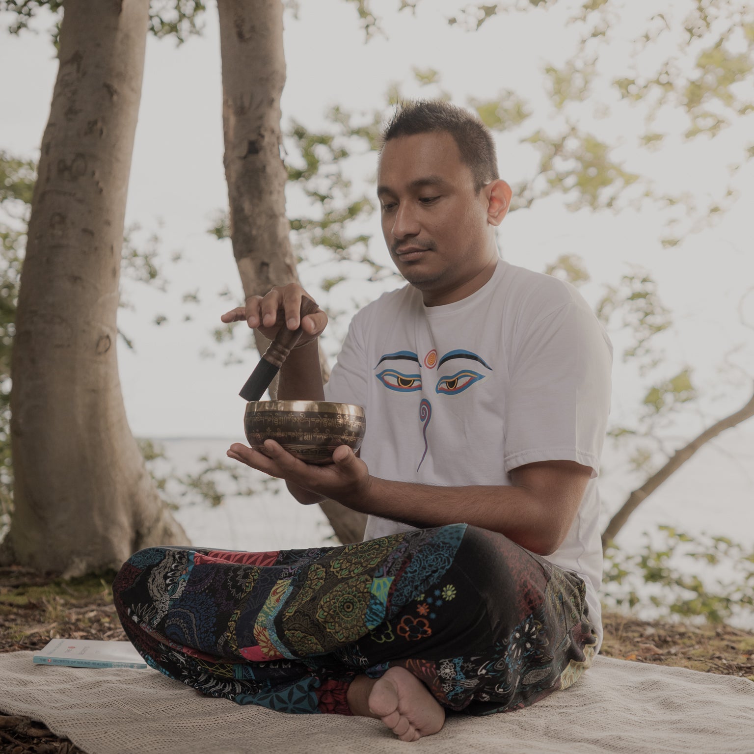 Person sitting outdoors holding a singing bowl with trees in the background