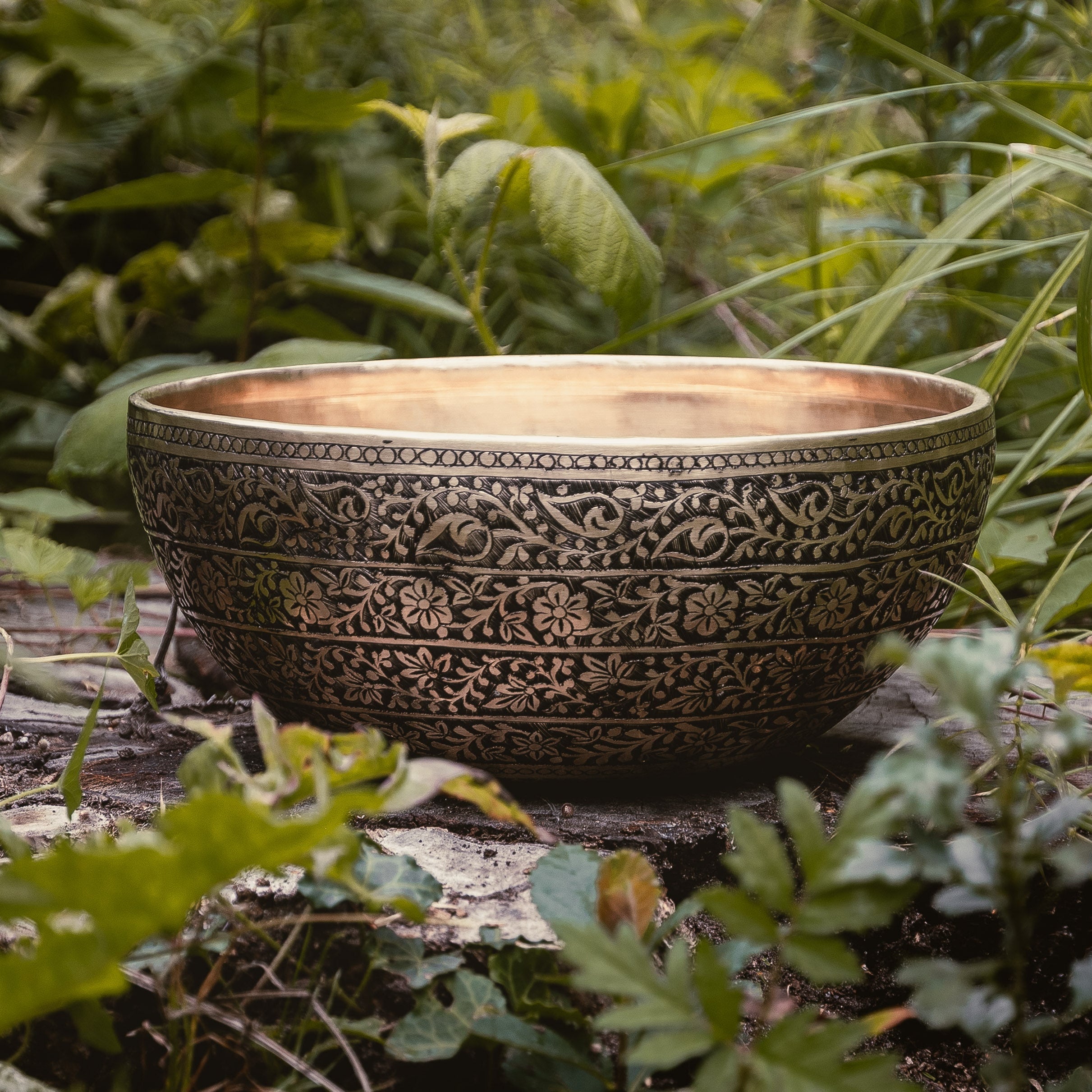 Decorative Singing bowl with intricate floral patterns on a wooden surface surrounded by greenery