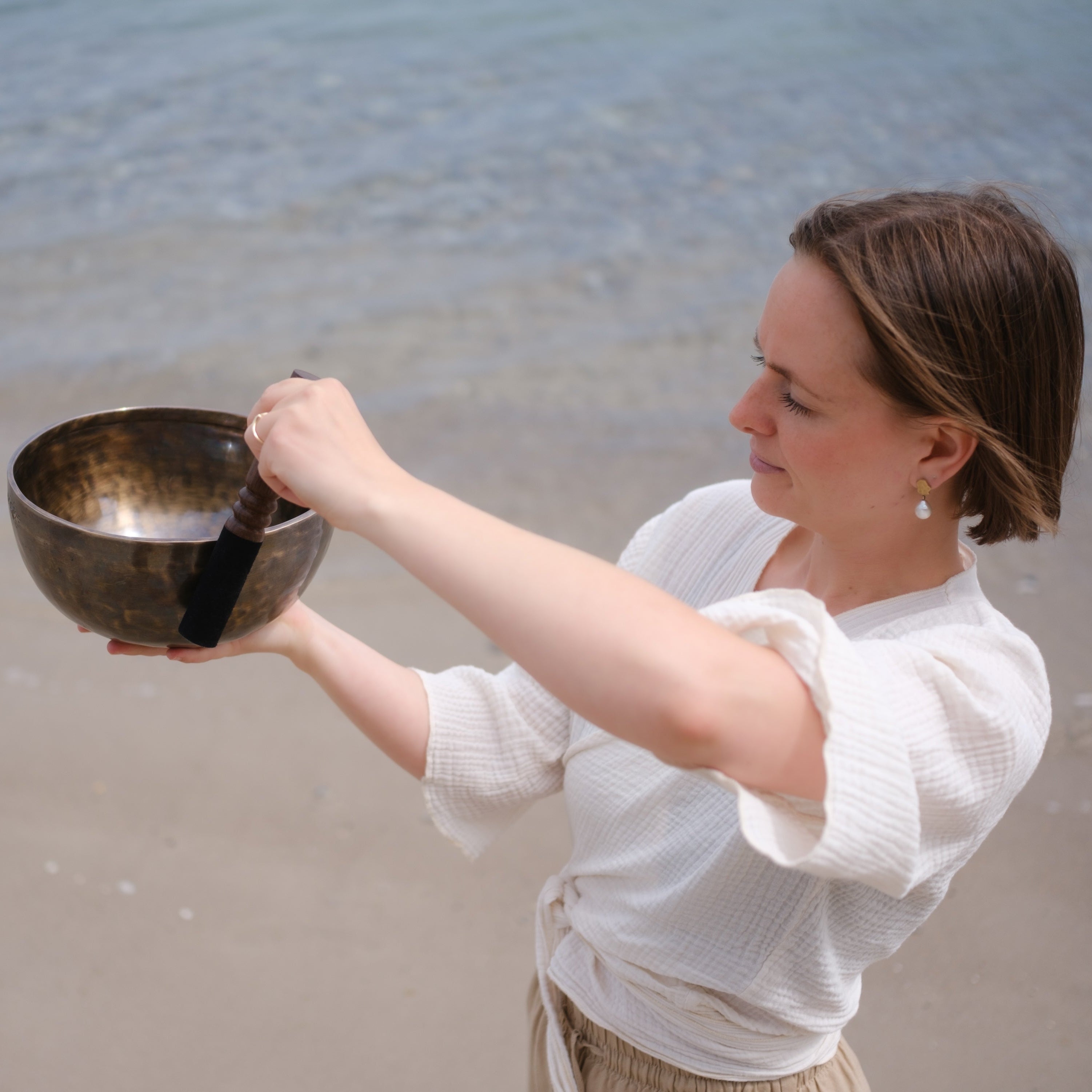 A woman standing on a sandy beach holds a singing bowl and striker, creating a sound. The background shows the ocean water and shoreline.