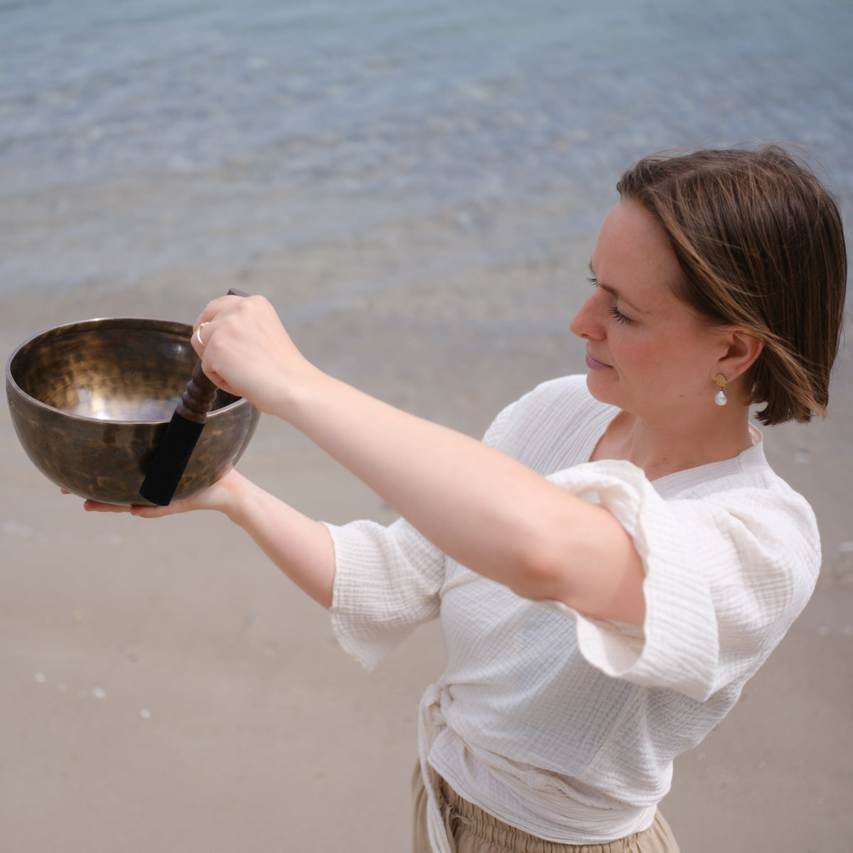En kvinde, der står på en sandstrand, holder en klangskål og en slagtang og frembringer en lyd. I baggrunden ses havet og kystlinjen.